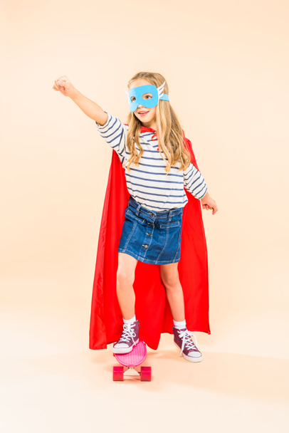 full length view of child in mask and hero cloak holding fist up while standing on skateboard on pink - Foto, imagen