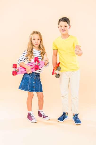 full length view of two smiling kids holding skateboards and showing thumbs up on pink - Photo, Image