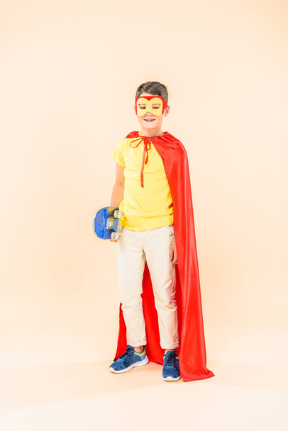 full length view of boy in mask and red cloak with skateboard on pink - Photo, image