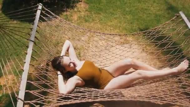 Młoda ładna kobieta w stroju pływakowego leżącego w hamaku wokół tropikalnych roślin i odpoczynku - Materiał filmowy, wideo