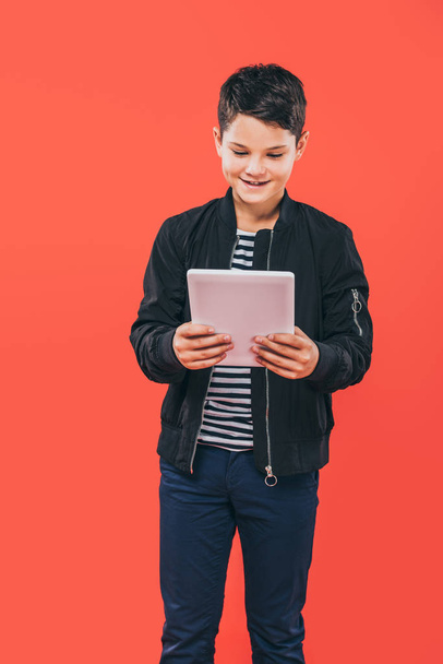 smiling kid in jacket using digital tablet isolated on red - Photo, Image