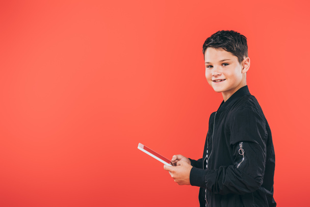 niño sonriente con chaqueta usando tableta digital aislada en rojo
 - Foto, imagen
