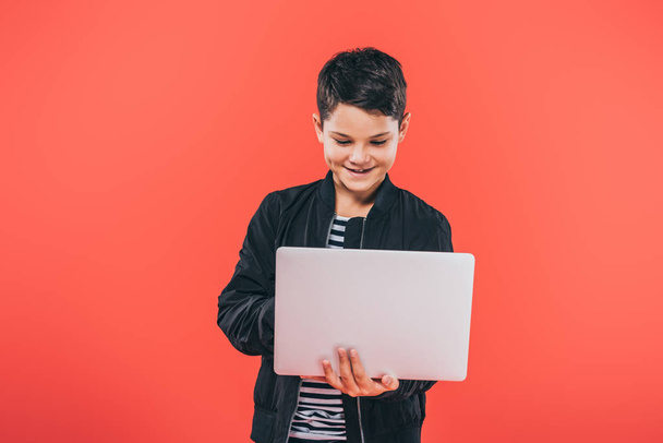 front view of smiling kid in jacket using laptop isolated on red - Foto, Imagem