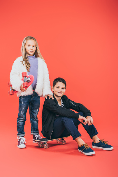 two smiling kids in autumn outfits with skateboards on red - Foto, afbeelding