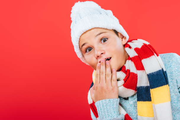 surprised kid in hat and scarf covering mouth with hand isolated on red - Foto, immagini