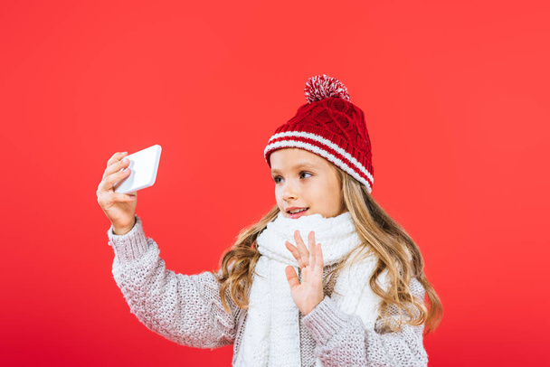 smiling kid in hat and scarf taking selfie isolated on red - Фото, изображение