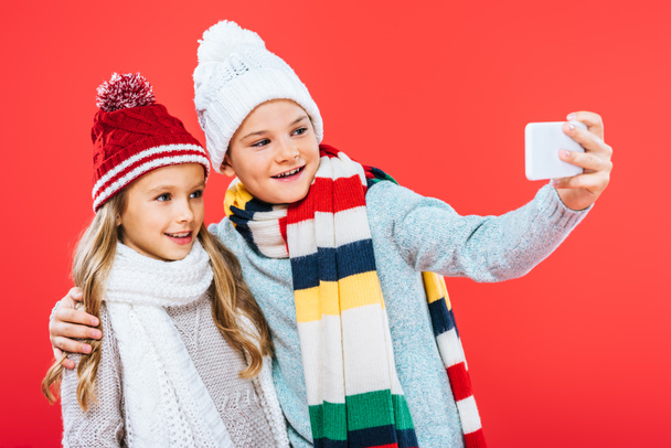 two smiling kids in winter outfits embracing and taking selfie isolated on red - Photo, Image