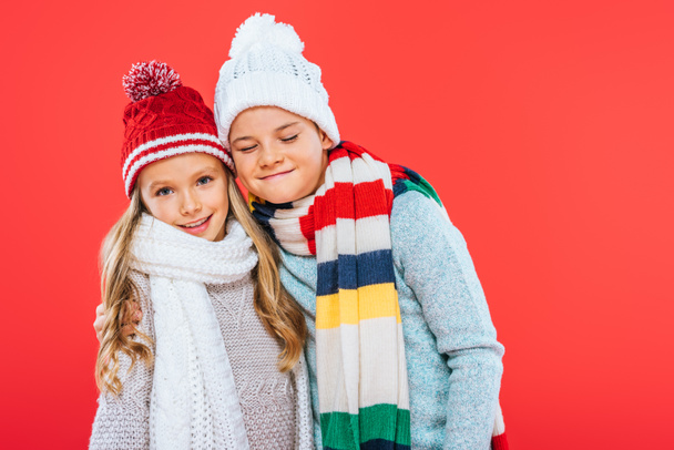 two smiling kids in hats and scarfs embracing isolated on red - Foto, afbeelding