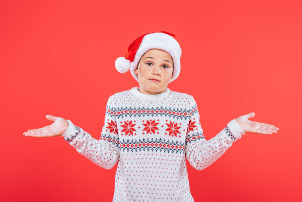 front view of child in sweater and santa hat showing shrug gesture isolated on red - Foto, afbeelding