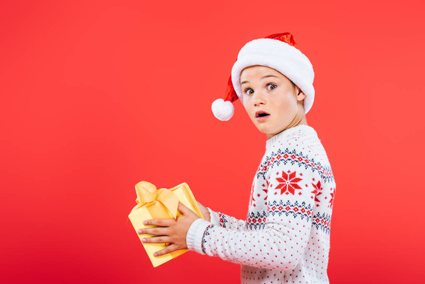 shocked kid in santa hat holding present isolated on red - Fotografie, Obrázek