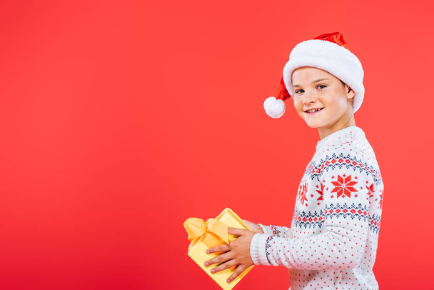 smiling kid in santa hat holding present isolated on red - Foto, afbeelding
