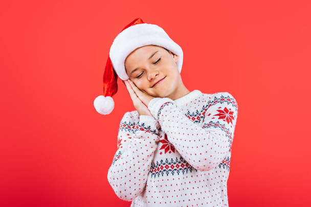 Kind in Pullover und Weihnachtsmütze schläft isoliert auf Rot - Foto, Bild