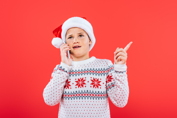 displeased child in santa hat talking on smartphone isolated on red - Fotó, kép