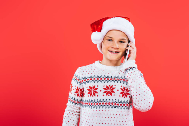 vista frontal de niño sonriente en sombrero de santa hablando en teléfono inteligente aislado en rojo
 - Foto, Imagen