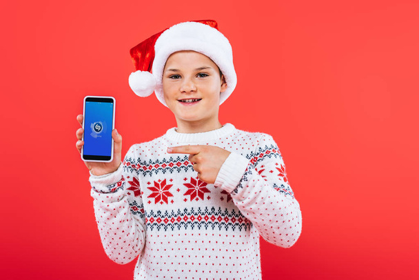 KYIV, UKRAINE - SEPTEMBER 9, 2019: front view of smiling kid in santa hat pointing with finger at smartphone with shazam app on screen - Foto, imagen
