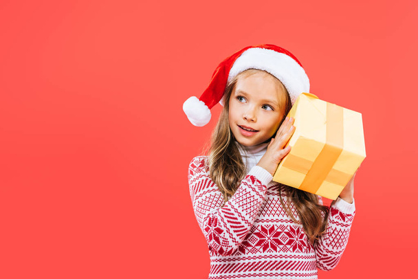 smiling kid in santa hat holding present isolated on red - Photo, Image
