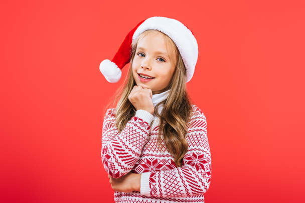 cute smiling kid in sweater and santa hat isolated on red - Photo, Image