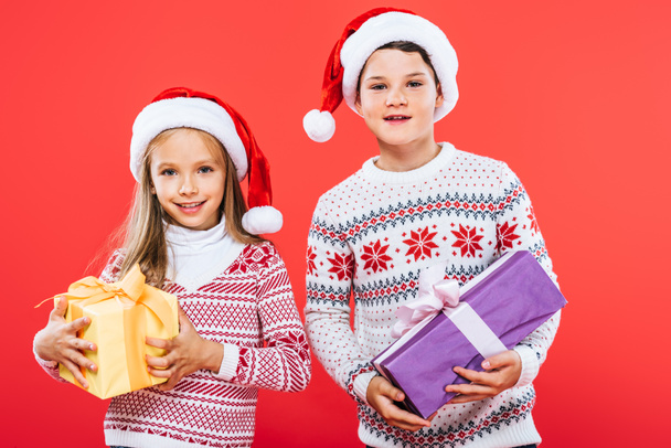 Vooraanzicht van twee lachende kinderen in Santa hoeden met cadeautjes geïsoleerd op rood - Foto, afbeelding