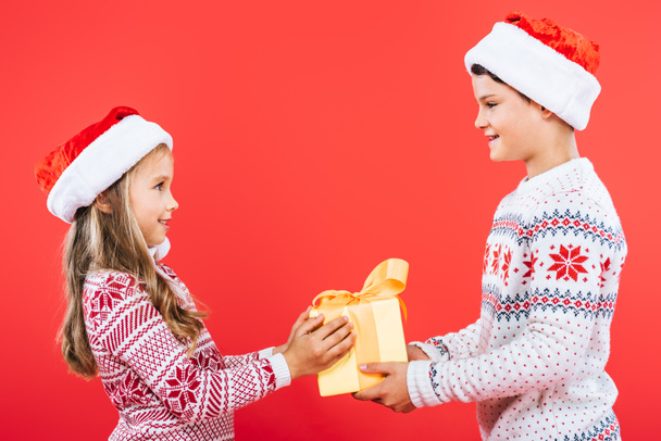 two smiling kids in santa hats with present looking at each other isolated on red - Foto, Imagem