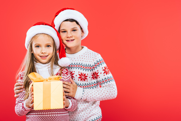 front view of smiling kids in santa hats with present embracing isolated on red - Photo, Image