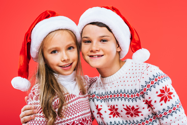 vue de face de deux enfants souriants dans des chapeaux de Père Noël et des pulls embrassant isolé sur rouge
 - Photo, image