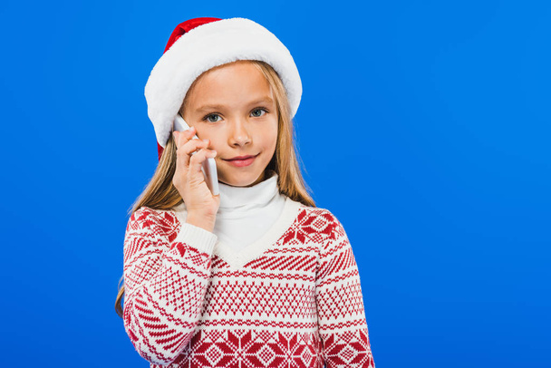 kid in santa hat and sweater talking on smartphone isolated on blue - Foto, imagen