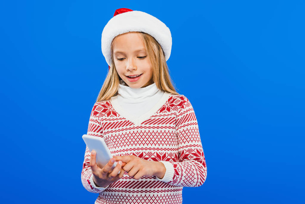 smiling kid in santa hat and sweater using smartphone isolated on blue - Photo, Image
