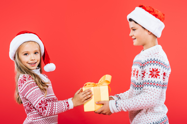 two smiling kids in santa hats with present isolated on red - Fotó, kép