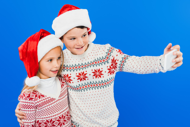 zwei lächelnde Kinder mit Weihnachtsmannmützen machen ein Selfie isoliert auf blauem Grund - Foto, Bild