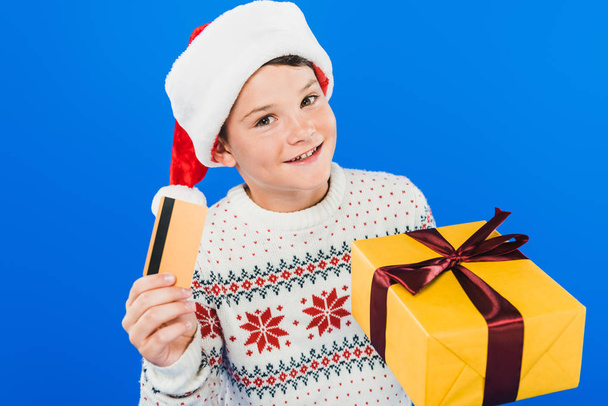 smiling kid in santa hat holding gift and credit card isolated on blue - Photo, image