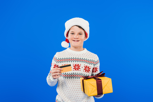 front view of smiling kid in santa hat holding gift and credit card isolated on blue - Фото, зображення