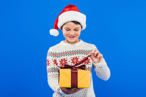 front view of kid in santa hat and sweater holding gift isolated on blue - Фото, изображение