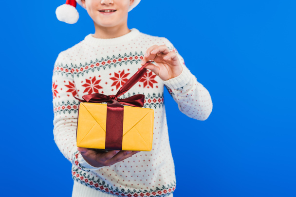 cropped view of kid in santa hat and sweater holding gift isolated on blue - 写真・画像