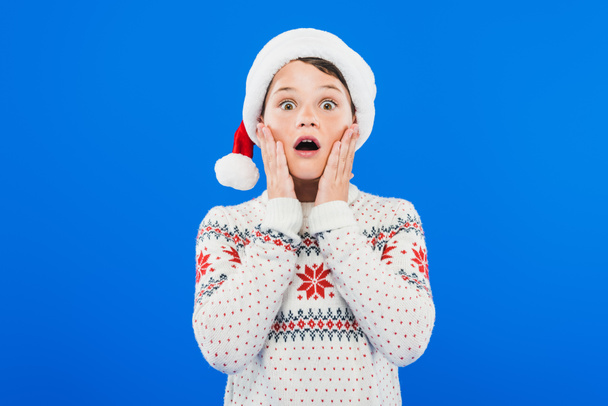 Frontansicht eines schockierten Kindes mit Weihnachtsmütze und Pullover isoliert auf blauem Grund - Foto, Bild