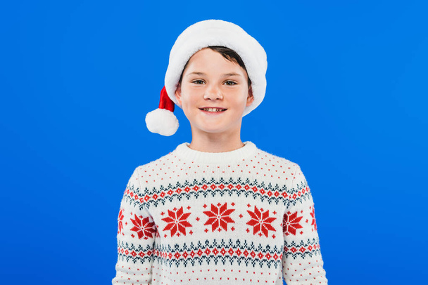 front view of smiling child in santa hat and sweater isolated on blue - Фото, зображення