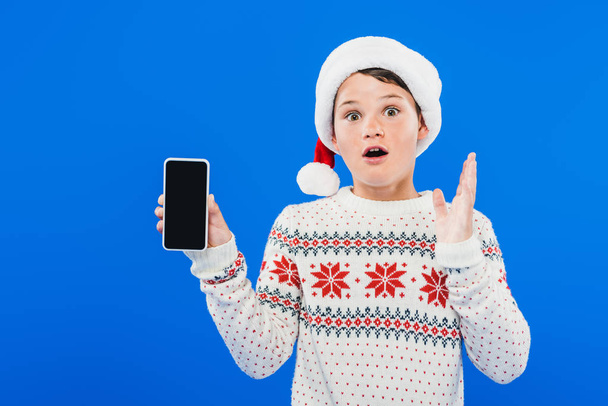 front view of shocked kid in santa hat holding smartphone with blank screen isolated on blue - Φωτογραφία, εικόνα