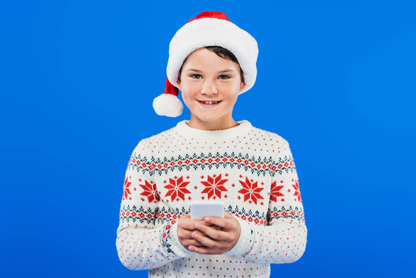 front view of smiling kid in santa hat using smartphone isolated on blue - Foto, Imagem