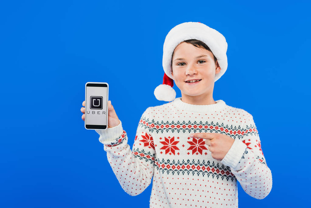 KYIV, UKRAINE - SEPTEMBER 9, 2019: front view of smiling kid in santa hat pointing with finger at smartphone with uber app on screen isolated on blue - Foto, Bild