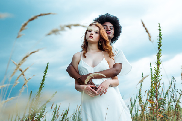 青空に赤毛の女の子を抱きしめるアフリカ系アメリカ人女性の選択的な焦点  - 写真・画像