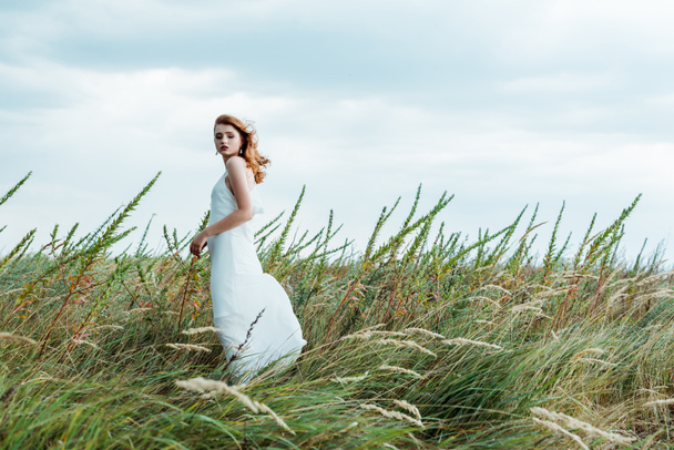 selective focus of attractive redhead woman in white dress looking at camera  - Photo, Image