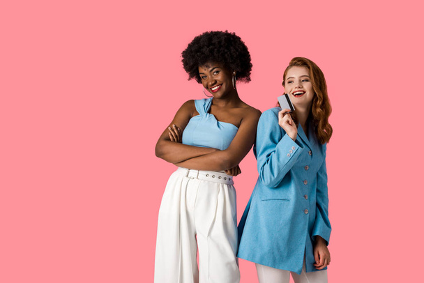 happy redhead girl holding credit card near african american woman standing with crossed arms isolated on pink  - Φωτογραφία, εικόνα