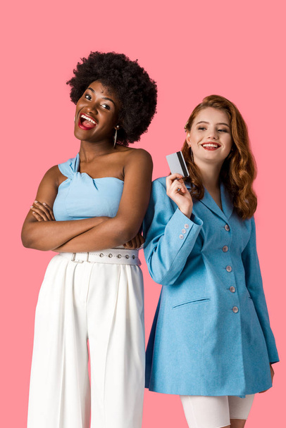 cheerful redhead girl holding credit card near african american woman standing with crossed arms isolated on pink  - Фото, зображення
