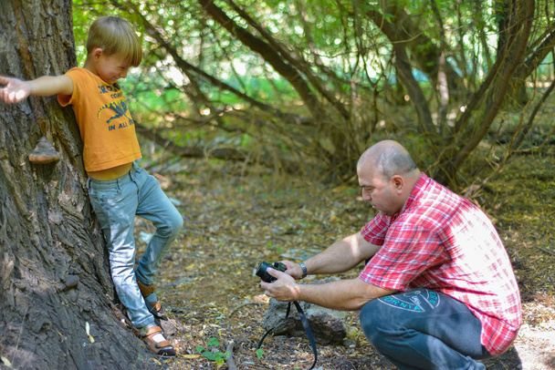 A man photographs a boy in a forest on nature against the background of a large tree. - Φωτογραφία, εικόνα