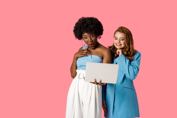 surprised african american woman looking at laptop with happy redhead girl isolated on pink  - Photo, Image