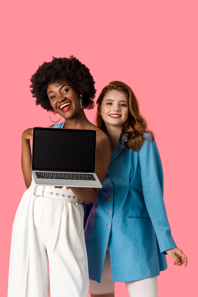 happy multicultural girls smiling near laptop with blank screen isolated on pink  - 写真・画像