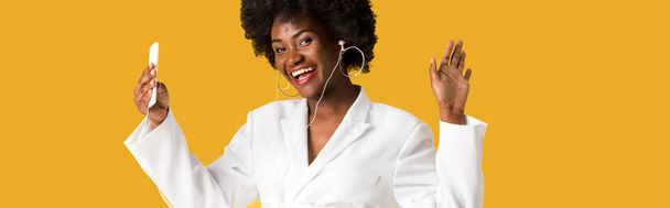panoramische shot van vrolijke Afro-Amerikaanse meisje luisteren muziek terwijl het houden van smartphone en gebaren geïsoleerd op oranje  - Foto, afbeelding