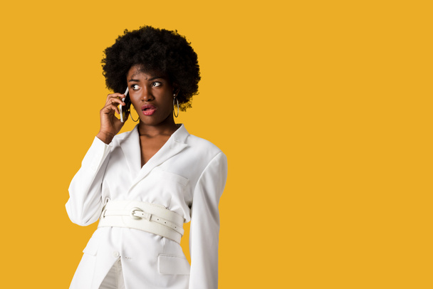 geschokt krullend Afro-Amerikaanse vrouw praten op smartphone geïsoleerd op oranje  - Foto, afbeelding