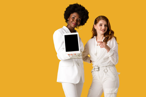 cheerful redhead girl pointing with finger near african american woman holding digital tablet with blank screen isolated on orange  - Fotografie, Obrázek