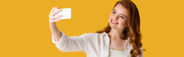 panoramiczny strzał szczęśliwy rudowłosy dziewczyna biorąc selfie izolowane na pomarańczowo  - Zdjęcie, obraz