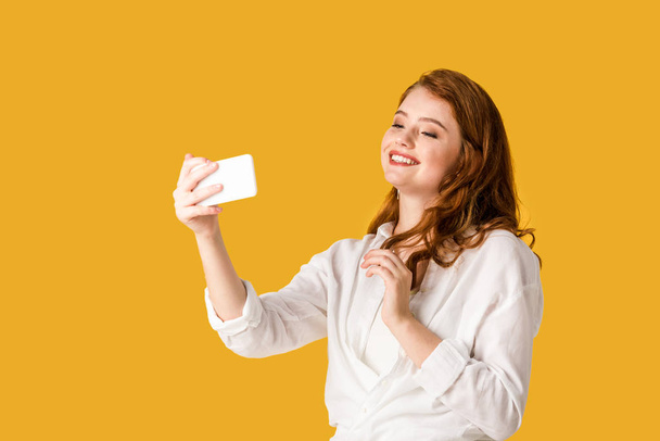 pretty redhead girl taking selfie and smiling isolated on orange  - Фото, зображення